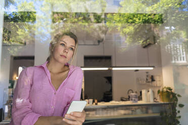 Nachdenklicher Jungunternehmer mit Smartphone durch Glas im Café gesehen - JOSEF14764