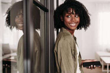 Smiling woman with smart phone leaning on glass door at home - EBBF06925