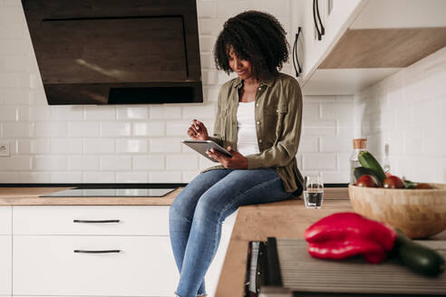 Woman using tablet PC sitting on kitchen counter at home - EBBF06950