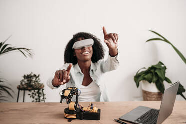 Smiling freelancer enjoying virtual reality headset with robotic model at home office - EBBF06981