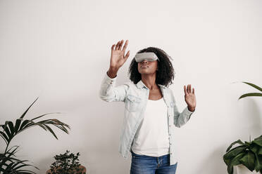 Woman wearing virtual reality headset standing in front of white wall at home - EBBF06983