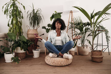 Woman practicing lotus position sitting on hassock by plants at home - EBBF06989