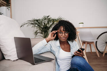 Businesswoman with smart phone and laptop on sofa at home - EBBF06995