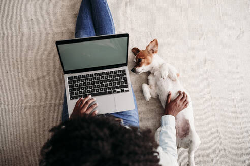 Frau streichelt Hund mit Laptop auf Teppich sitzend zu Hause - EBBF06999