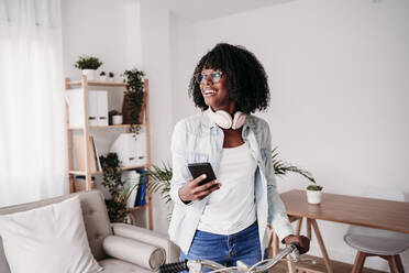 Contemplative woman holding smart phone with bicycle at home - EBBF07006