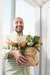 Lächelnder Mann mit frischem Bio-Gemüse in einer Papiertüte in der Nähe der Tür - KMKF01885