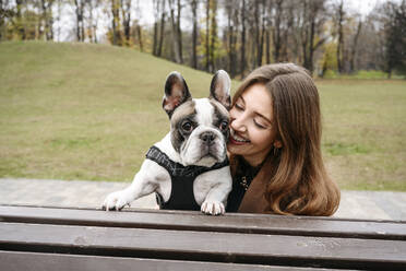 Cheerful woman embracing French bulldog on bench at park - EYAF02344