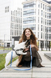 Happy woman playing with French bulldog on footpath - EYAF02347
