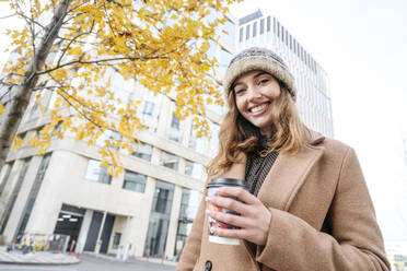 Glückliche Frau mit Kaffeetasse vor einem Gebäude stehend - EYAF02354