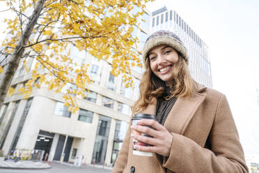 Happy woman with coffee cup standing in front of building - EYAF02354