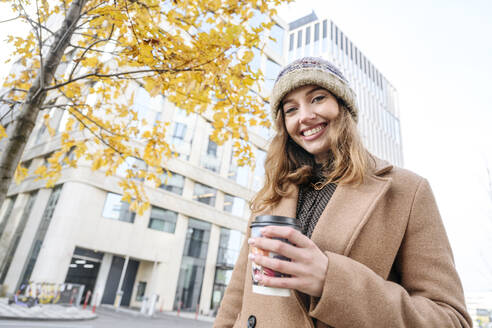 Glückliche Frau mit Kaffeetasse vor einem Gebäude stehend - EYAF02354