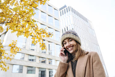 Glückliche Frau mit Strickmütze, die vor einem Gebäude mit ihrem Smartphone telefoniert - EYAF02355