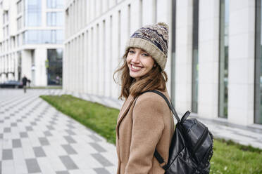 Happy woman wearing knit hat standing with backpack on footpath - EYAF02363