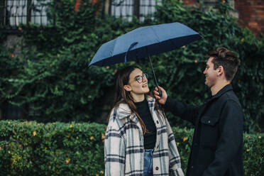Junger Mann gibt seiner Freundin vor Pflanzen einen Regenschirm - VSNF00101