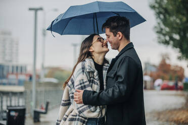 Lächelnder junger Mann und Frau küssen sich unter einem Regenschirm auf einem Fußweg - VSNF00103
