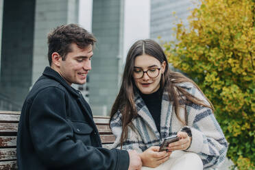 Girlfriend sharing mobile phone with boyfriend on bench - VSNF00104