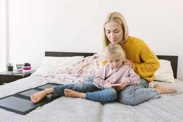 Daughter sitting with mother using smart phone on bed at home - SIF00532
