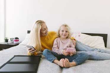 Smiling mother and daughter sitting with smart phones on bed in bedroom - SIF00533