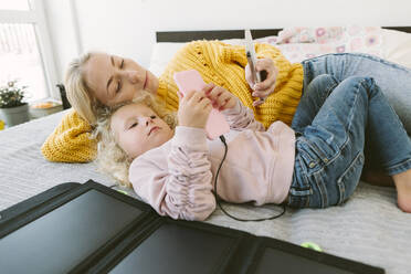 Mother and daughter using mobile phones lying on bed at home - SIF00535