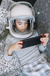 Niedliches Kind im Schlafanzug auf der Couch liegend, während es zu Hause mit einer Videospielkonsole spielt - ADSF40311