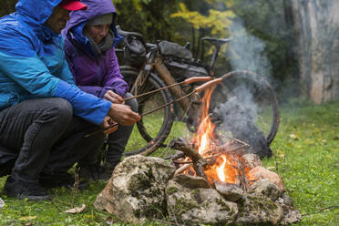 Seitenansicht von Radfahrern, die sich an einer Feuerstelle wärmen und Würstchen am Stock zubereiten, auf einer grünen Wiese an einem kalten Tag - ADSF40332