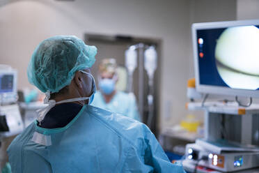 Back view of man in surgeon uniform looking at monitor while performing surgery on blurred background of modern operating theater - ADSF40341