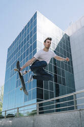 Von unten junger Mann mit Beinprothese springt auf Skateboard über dem Boden gegen modernes Gebäude an sonnigem Tag - ADSF40358