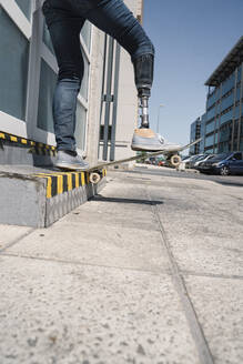 Von unten junger Mann mit Beinprothese springt auf Skateboard über dem Boden gegen modernes Gebäude an sonnigem Tag - ADSF40360