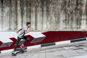 Side view of young guy with leg prosthesis running up paved slope against shabby building during workout on city street - ADSF40361