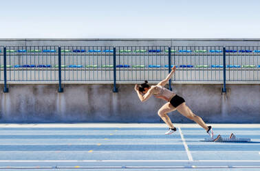 Side view of young strong sportswoman beginning to run fast from starting blocks during track and field workout on stadium - ADSF40370