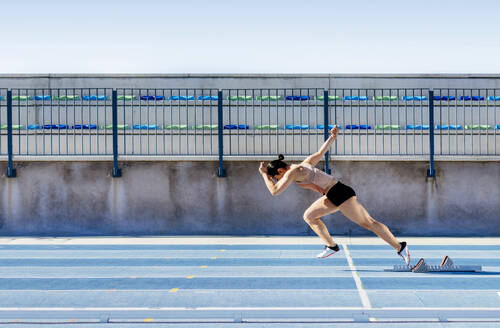 Seitenansicht einer jungen, starken Sportlerin, die während eines Leichtathletik-Trainings im Stadion schnell aus den Startblöcken zu laufen beginnt - ADSF40370