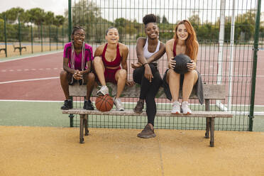 Lächelnde junge Freunde sitzen auf einer Bank am Basketballplatz - JCCMF07886