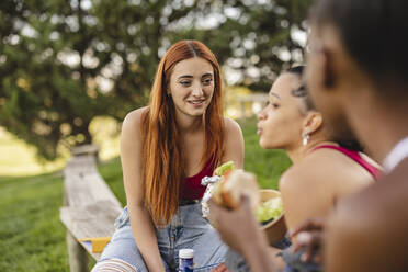 Junge Frau sieht ihren Freund an, der mit Essen im Park sitzt - JCCMF07923