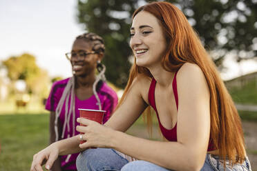 Happy redhead woman sitting with drink by friend at park - JCCMF07937