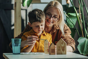 Happy mother and son painting wooden model houses at table - VSNF00131