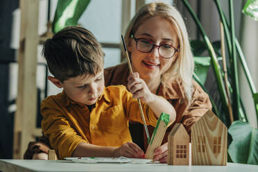 Smiling mother and son painting wooden model houses at table - VSNF00132