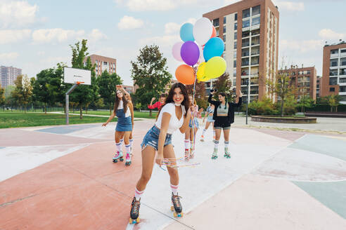 Smiling young woman wearing roller skates holding colorful balloons in front of friends at sports court - MEUF08474