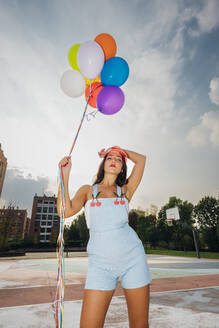 Young woman holding colorful balloons at sports court - MEUF08491