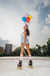 Smiling woman wearing roller skates holding colorful balloons at sports court - MEUF08494