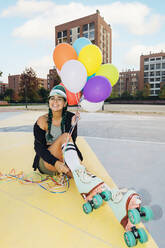 Glückliche Frau auf Rollschuhen mit bunten Luftballons auf dem Sportplatz - MEUF08517