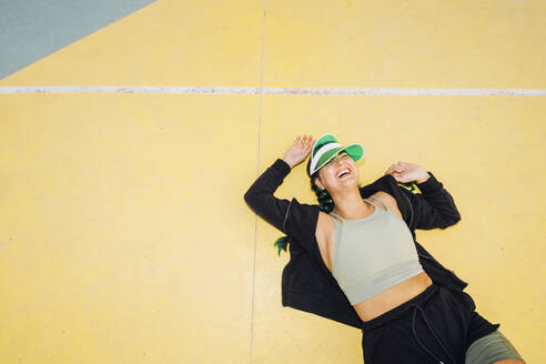 Happy young woman wearing sun visor lying on sports court - MEUF08520