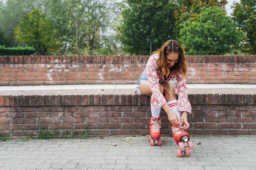 Smiling woman lacing up roller skate on wall - MEUF08561