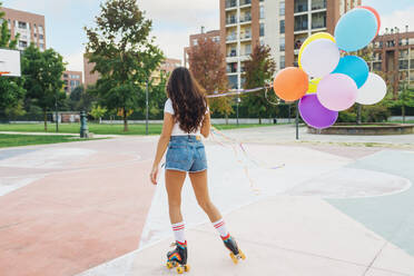 Young woman holding colorful balloons roller skating at sports court - MEUF08571