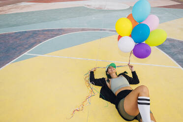 Happy woman holding colorful balloons lying on sports court - MEUF08575