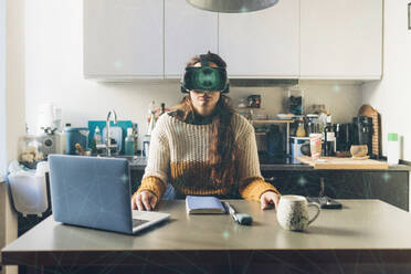 Frau mit Virtual-Reality-Brille sitzt vor einem Laptop im Home-Office - MEUF08582