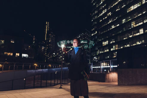 Woman using augmented reality standing on footpath at night - MEUF08588