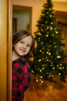 Lächelnder Junge, der zur Tür hereinschaut, mit beleuchtetem Weihnachtsbaum im Hintergrund - ANAF00506