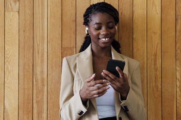 Happy businesswoman using mobile phone in front of wooden wall - PGF01330