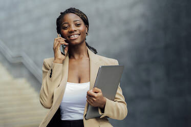 Happy businesswoman talking on mobile phone holding laptop - PGF01345