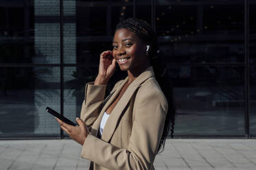 Lächelnde Geschäftsfrau mit Smartphone und drahtlosen In-Ear-Kopfhörern auf dem Fußweg - PGF01375